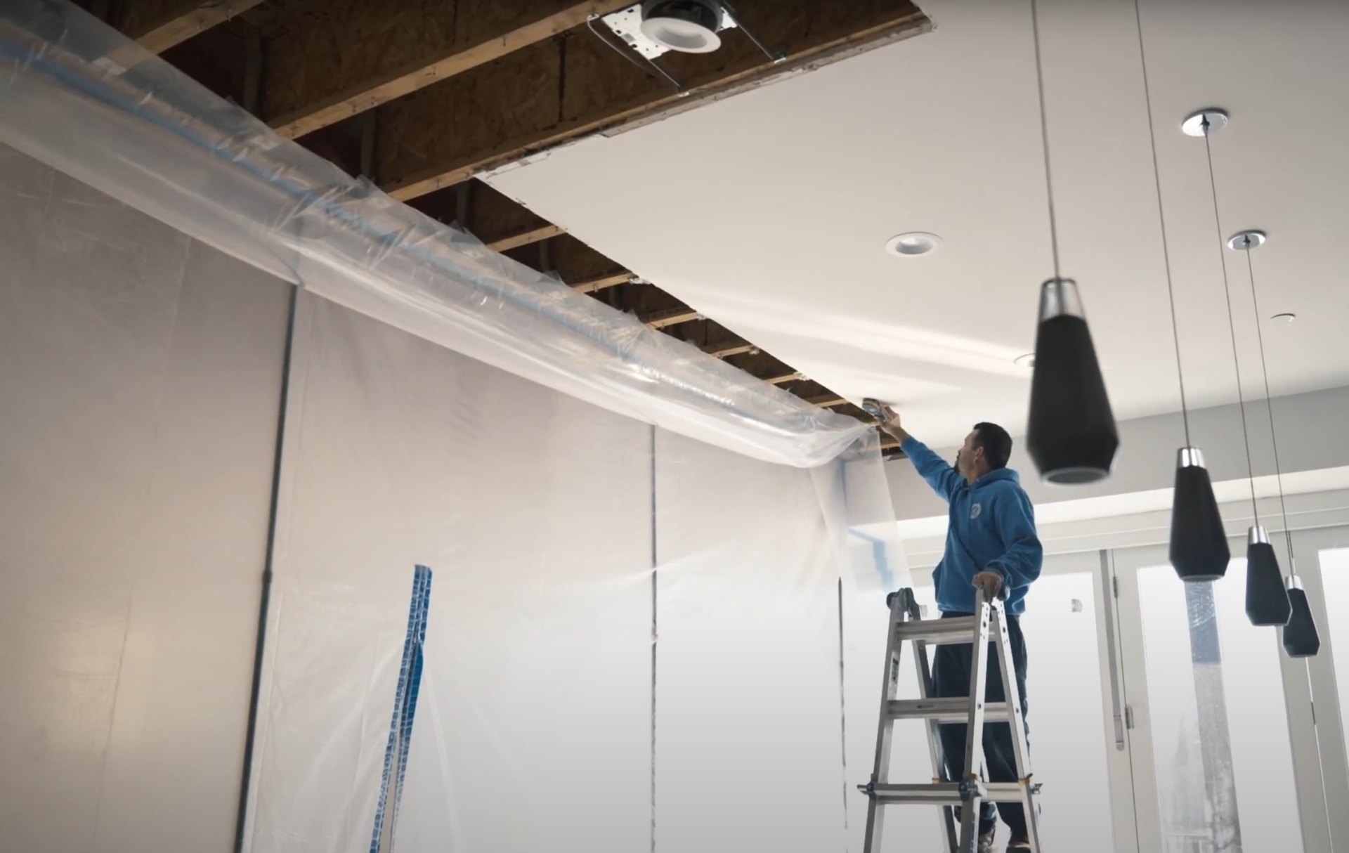 Professional water damage restoration work on commercial building systems and ceiling infrastructure Water damage restoration technician working on exposed ceiling and HVAC systems with protective plastic sheeting
